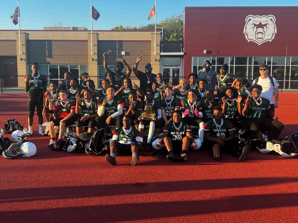 A football team poses on a track with helmets, medals and a trophy.